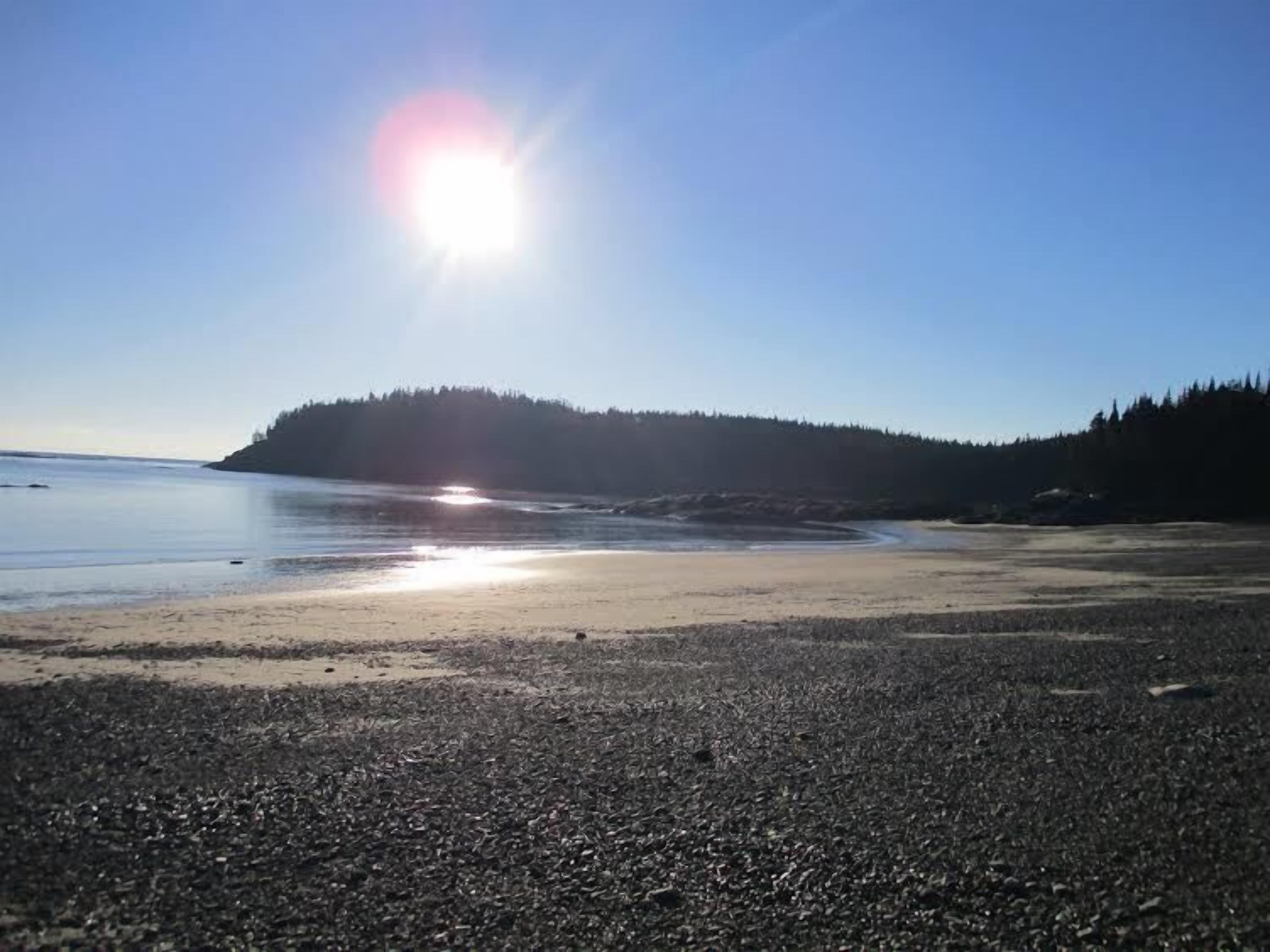 A beach with pebbles, calm water, and a forested hill in the background.