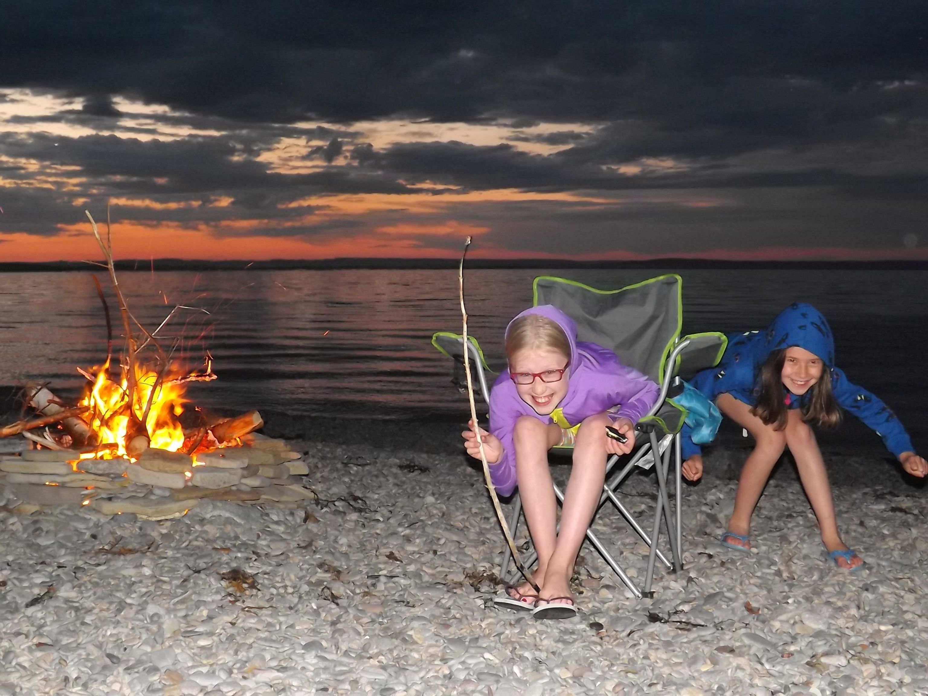 Deux personnes assises sur des chaises près d’un feu de camp sur une plage de galets, au coucher du soleil.