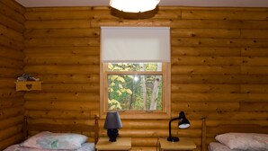 Quarto de cabana de toras com duas camas, uma escrivaninha com luminárias e uma janela com vista para árvores.