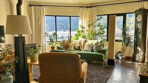 A living room with a green sofa, multiple potted plants, a lamp, and a view of a building through large windows. - Walk everywhere from Railtown Suite! + spacious, beautiful, & comfortable. (Nelson)