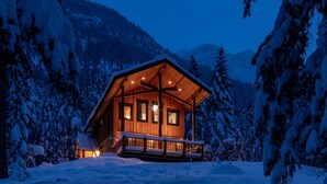 Eine Holzjagdhaus mit Veranda, umgeben von schneebedeckten Bäumen und Bergen.