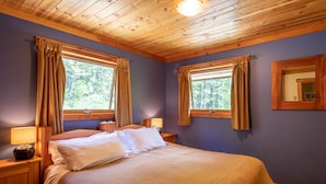 Ein Schlafzimmer mit Holzboden, zwei Fenstern mit Vorhängen, einem Bett mit weißer Bettwäsche und einem Nachttisch mit Lampe.