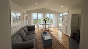 Un vaste espace de vie comprenant un canapé, une table à manger avec des chaises, une vue sur la mer grâce à de grandes fenêtres, et un coin cuisine avec des placards.