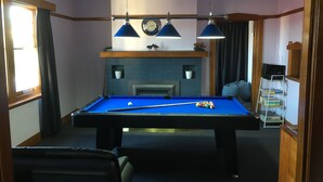 Una mesa de billar con superficie azul de fieltro, rodeada de sillas, en una habitación con chimenea, un reloj y una estantería.