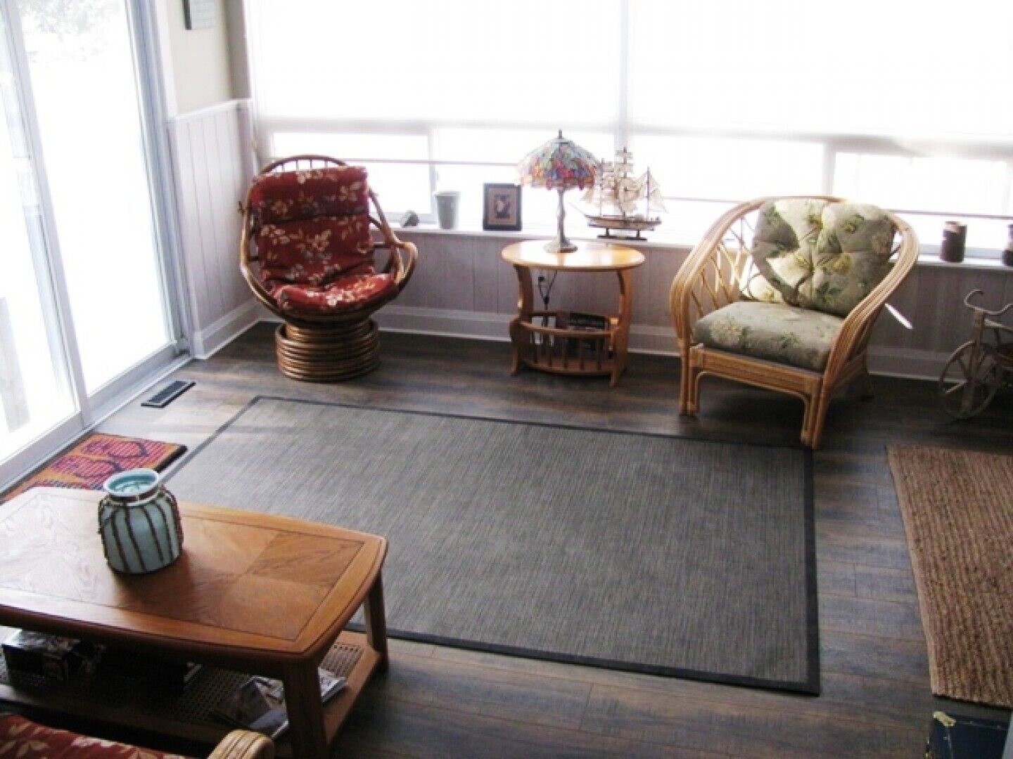 Una habitación acogedora con un ventanal grande, una silla de mimbre, una mesita y una lámpara decorativa.