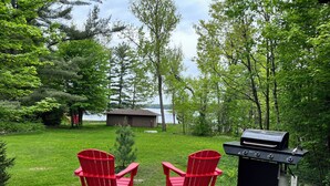 A deck with two red Adirondack chairs facing a lake, a grill, and a small building in the background.