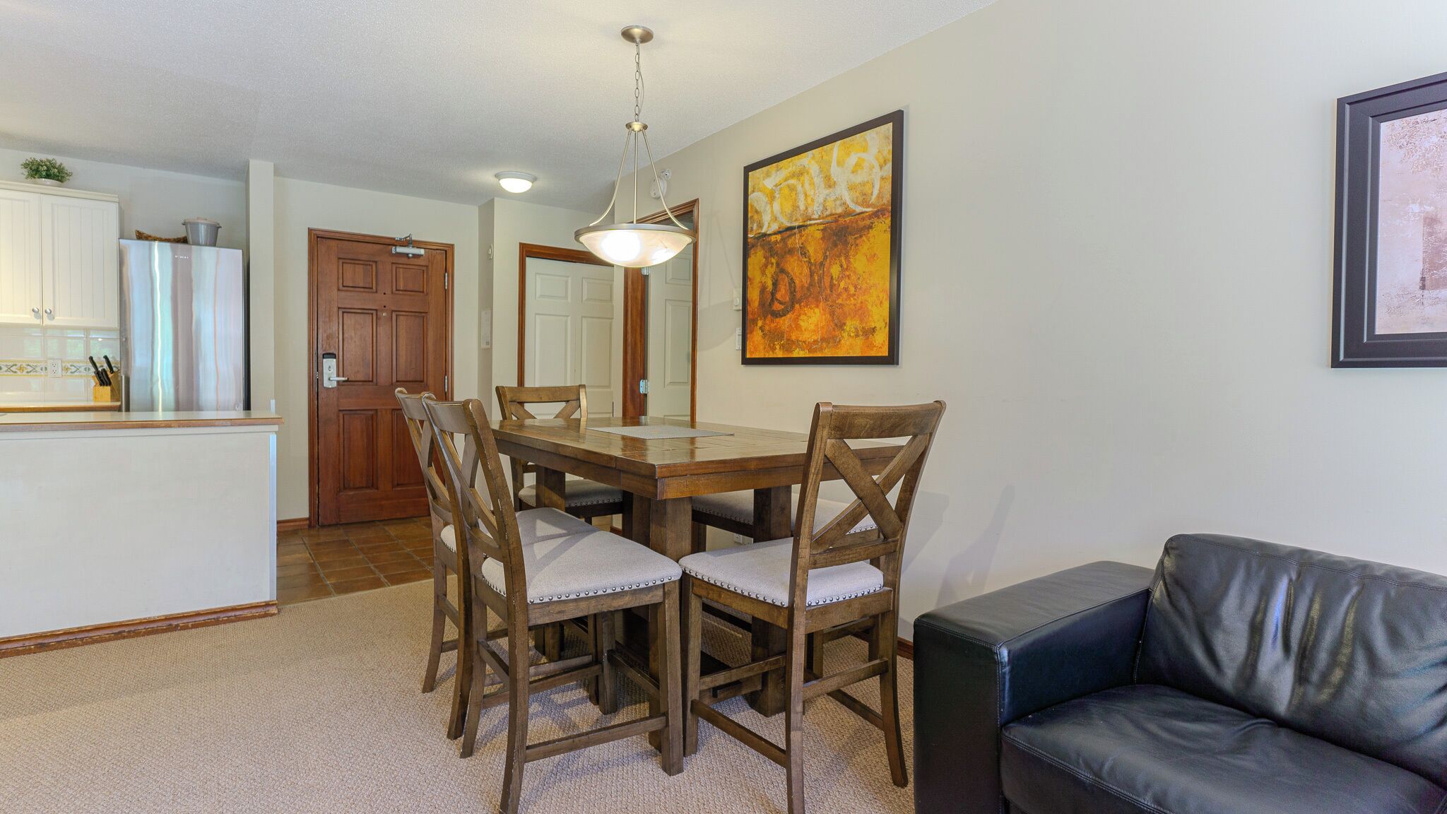 A dining area with a wooden table and chairs, a kitchen in the background, and a black leather sofa.