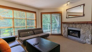 A living room with a stone fireplace, a sofa, a coffee table, and large windows with blinds. - Aspens #549 Prime Ski-in Ski-out Location! Top Floor Unit, Pool, Hot tubs, BBQ (Whistler)