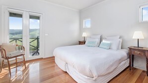 Un dormitorio con una cama grande, dos mesitas de noche con lámparas, una silla de madera y un ventanal corredero que da a un balcón con vistas al campo.