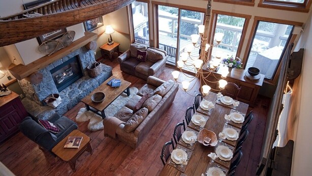 Amplia sala de estar con chimenea de piedra, sofás de cuero, una mesa de comedor preparada para seis personas y un ventanal con vistas a un paisaje nevado.
