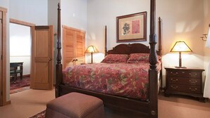 Una cama con dosel con colcha estampada, dos mesitas de noche con lámparas, un aparador de madera y una puerta que da a otra habitación.