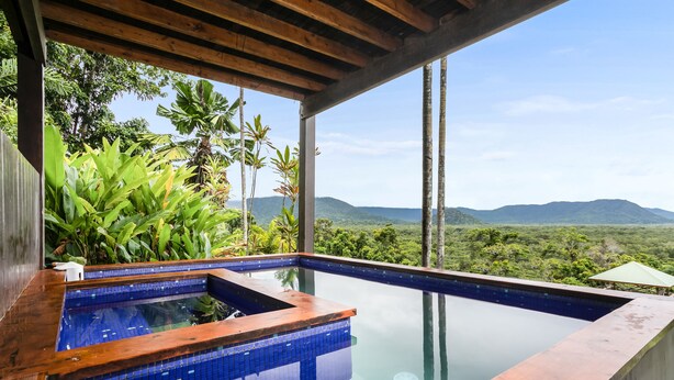 Ein moderner Infinity-Pool mit Holzterrasse, umgeben von üppiger Vegetation und Blick auf ferne Berge.