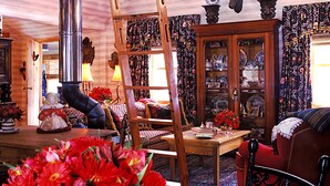 Um interior aconchegante com teto de madeira, lareira, área de refeições com mesa e cadeiras, e sala de estar com sofá e lustre decorativo em forma de chifres.