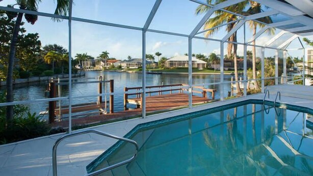 A pool with a glass enclosure, a wooden deck, and a view of a lake and houses in the background. - 3/2 Home steps away from Tiger Tail access (Marco Island)