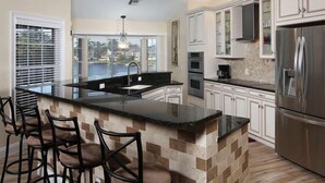 A modern kitchen with a large island, bar stools, stainless steel appliances, and a view of the outdoors through large windows. - 3/2 Home steps away from Tiger Tail access (Marco Island)