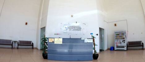 A reception desk with a sign, two benches, a vending machine, and potted plants in a spacious lobby with tiled flooring.