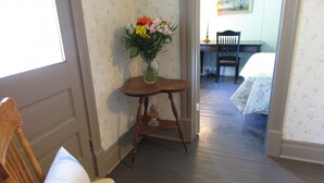 Una silla de madera con cojín, una pequeña mesa de madera con un jarrón de flores, y una cama con colcha estampada en una habitación con papel tapiz floral y suelo de madera.