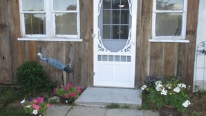 Una casa de madera con puerta blanca, dos ventanas y un pequeño jardín con plantas en macetas y flores.