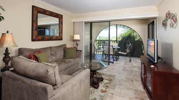 A living room with a sofa, coffee table, and a TV on a wooden cabinet. There's a balcony with a table and chairs. - Lovely condo on the Bay with inland view! (Marco Island)