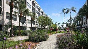 A pathway flanked by palm trees and purple flowers leading to a building with a modern architectural style. - Lovely condo on the Bay with inland view! (Marco Island)