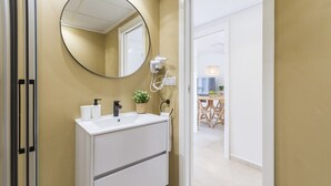 Een badkamer met een wit wastafelmeubel, een ronde spiegel, een haardroger en een potplant.