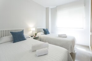 Quarto de hotel com duas camas, roupa de cama branca, mesa de cabeceira com abajur e uma janela.