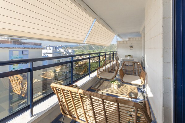 A balcony with wicker furniture, a glass railing, and a view of buildings and greenery. - Amazing modern style condo, wifi free, interantional channels, heating swimming pool, children's area, BBQ - only families (Playa de Gandía)