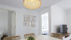 Um quarto com uma mesa branca, cadeiras de vime, uma TV sobre um suporte de madeira e quadros nas paredes.