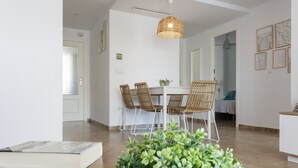 Uma área de refeições com mesa branca, cadeiras de vime, luminária pendente e um vaso com planta.