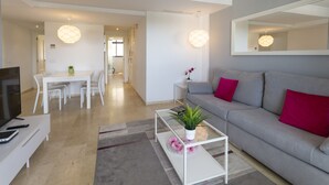 Een moderne woonkamer met een grijze bank, een salontafel met een plant, een eethoek met een witte tafel en stoelen, en een gang met een deur naar een andere kamer.