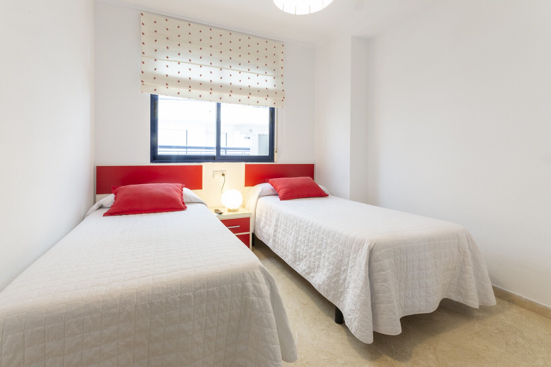 A double room with two beds, each with a red pillow, a bedside table with a lamp, and a window with a polka dot blind.