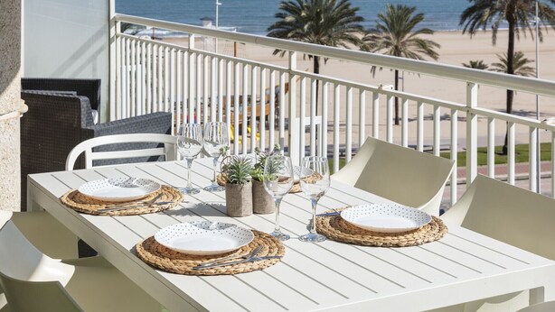 A balcony with a white table set for two, overlooking a beach with palm trees and the ocean. - Apartment with amazing Ocean Views / Pool - Only families (Playa de Gandía)