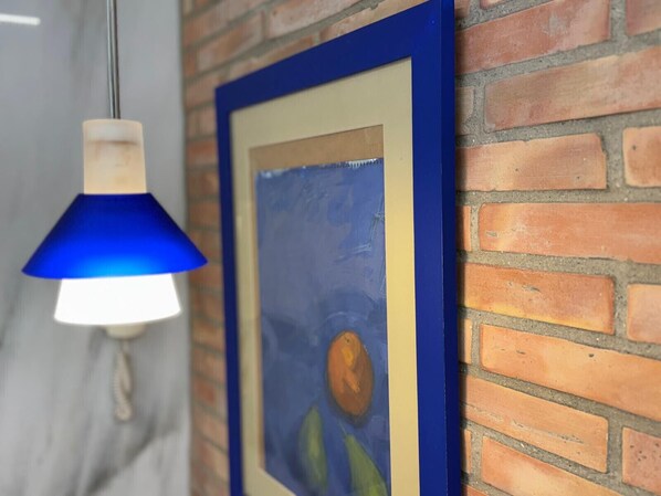 Une œuvre d’art encadrée accrochée à un mur de briques, un luminaire suspendu bleu et une vue partielle d’une pièce dont le sol est gris.