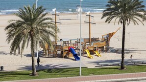 A beach playground with slides and a wooden structure, palm trees, and a clear view of the ocean. - Apartment with amazing Ocean Views / Pool - Only families (Playa de Gandía)