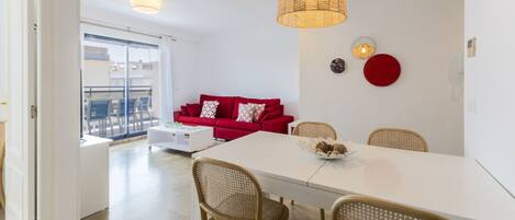 A modern living room with a white sofa, red cushions, a dining table with wicker chairs, and a hanging light fixture.