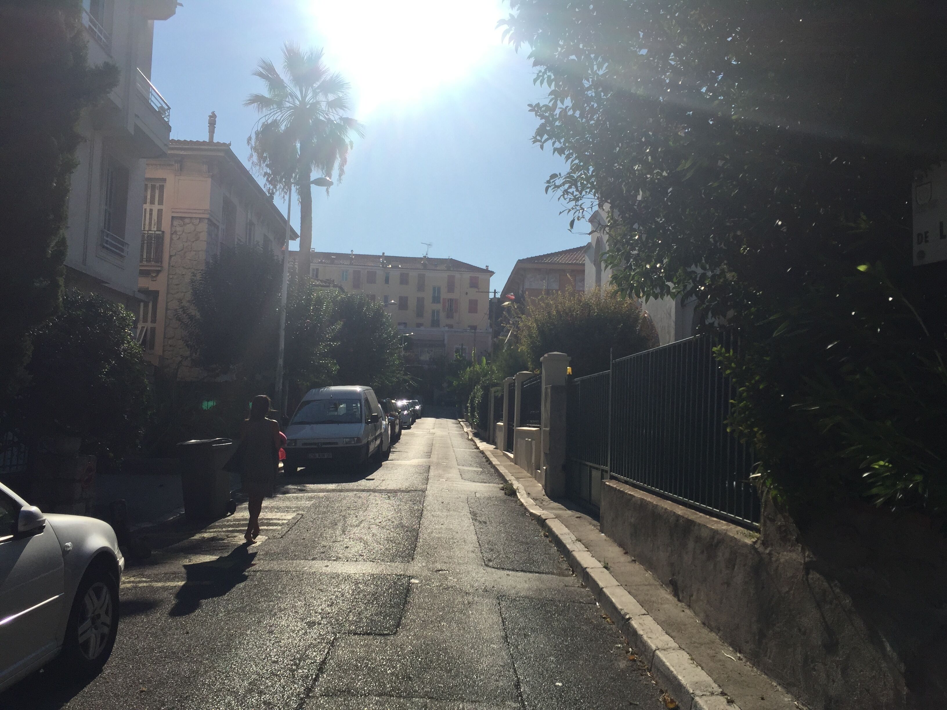 A narrow urban street with parked cars, a pedestrian, and buildings on either side.