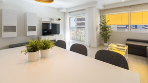 Un salon moderne comprenant un coin repas, un téléviseur à écran plat fixé au mur, une grande fenêtre avec des rideaux et un balcon aménagé avec du mobilier d’extérieur.