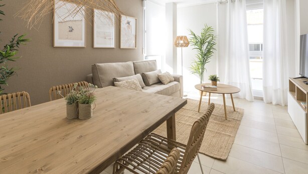 A modern living room with a wooden dining table, a sofa, and a TV. There are plants and artwork on the walls.
