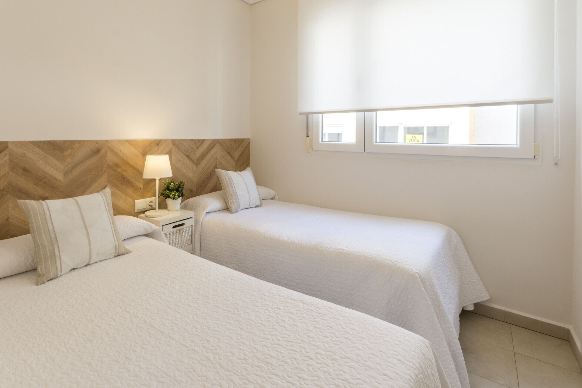 Et hotelværelse med to senge, et natbord med lampe, en potteplante og et vindue med udsigt til bygninger.