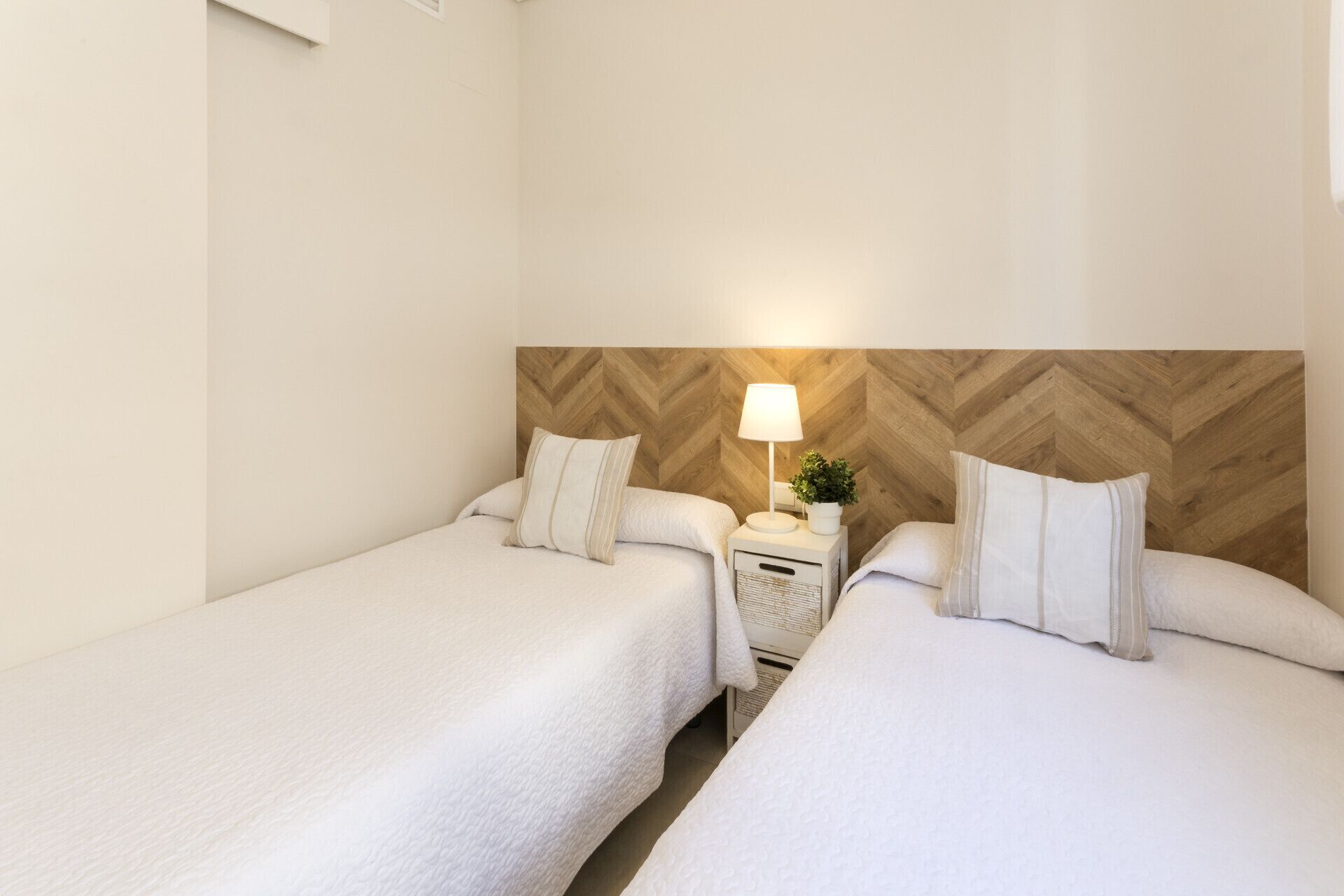 Quarto de hotel com duas camas de solteiro, cabeceira de madeira, mesa de cabeceira com luminária e uma pequena planta em vaso.