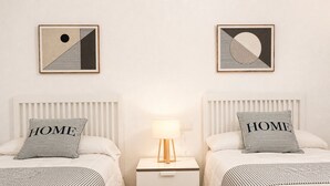 Ein Hotelzimmer mit zwei Betten, jedes mit einem Kopfkissen mit dem Wort 'HOME', einem Nachttisch mit einer Lampe und zwei gerahmten abstrakten Kunstwerken an der Wand.