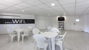 Ein Konferenzraum mit weißen Kunststoffstühlen und -tischen, einem großen Schild mit der Aufschrift "ZONA WiFi", einem Fernseher auf einem Standfuß und einer Tür mit rundem Fenster.