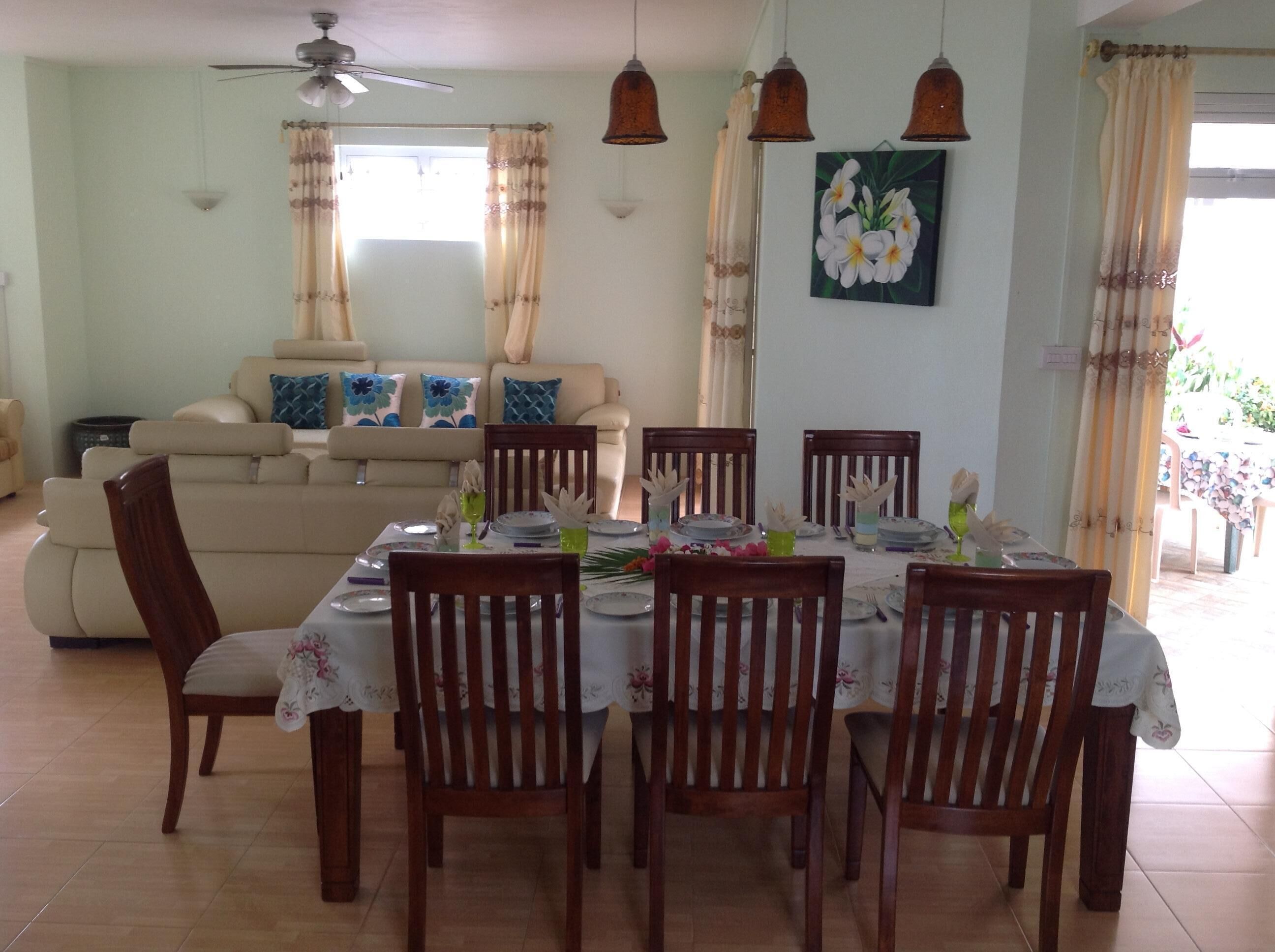 A dining area with a table set for six, surrounded by wooden chairs. There is a ceiling fan above and a dining table with a floral tablecloth.