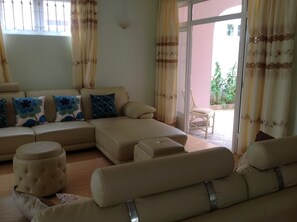 Un salon avec un canapé d’angle beige, un pouf rond et une chaise longue. Une chaise est placée à l’extérieur de la porte-fenêtre coulissante.