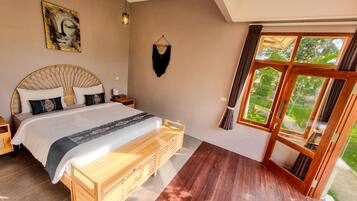 A bedroom with a large bed, wooden headboard, bedside tables, a bench, a wall-mounted artwork, and a large window with curtains.