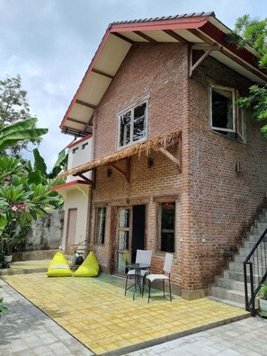 A two-story brick house with a red roof, a staircase, and a patio with chairs and cushions. - Les Rizieres Lombok (Tetebatu)