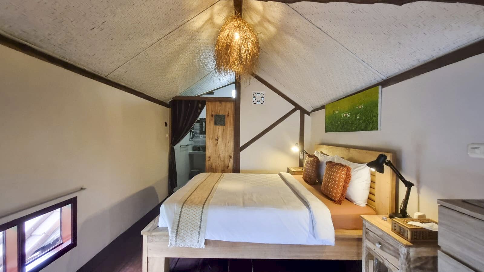 A bedroom with a bed, bedside table, lamp, and a view of the outside.