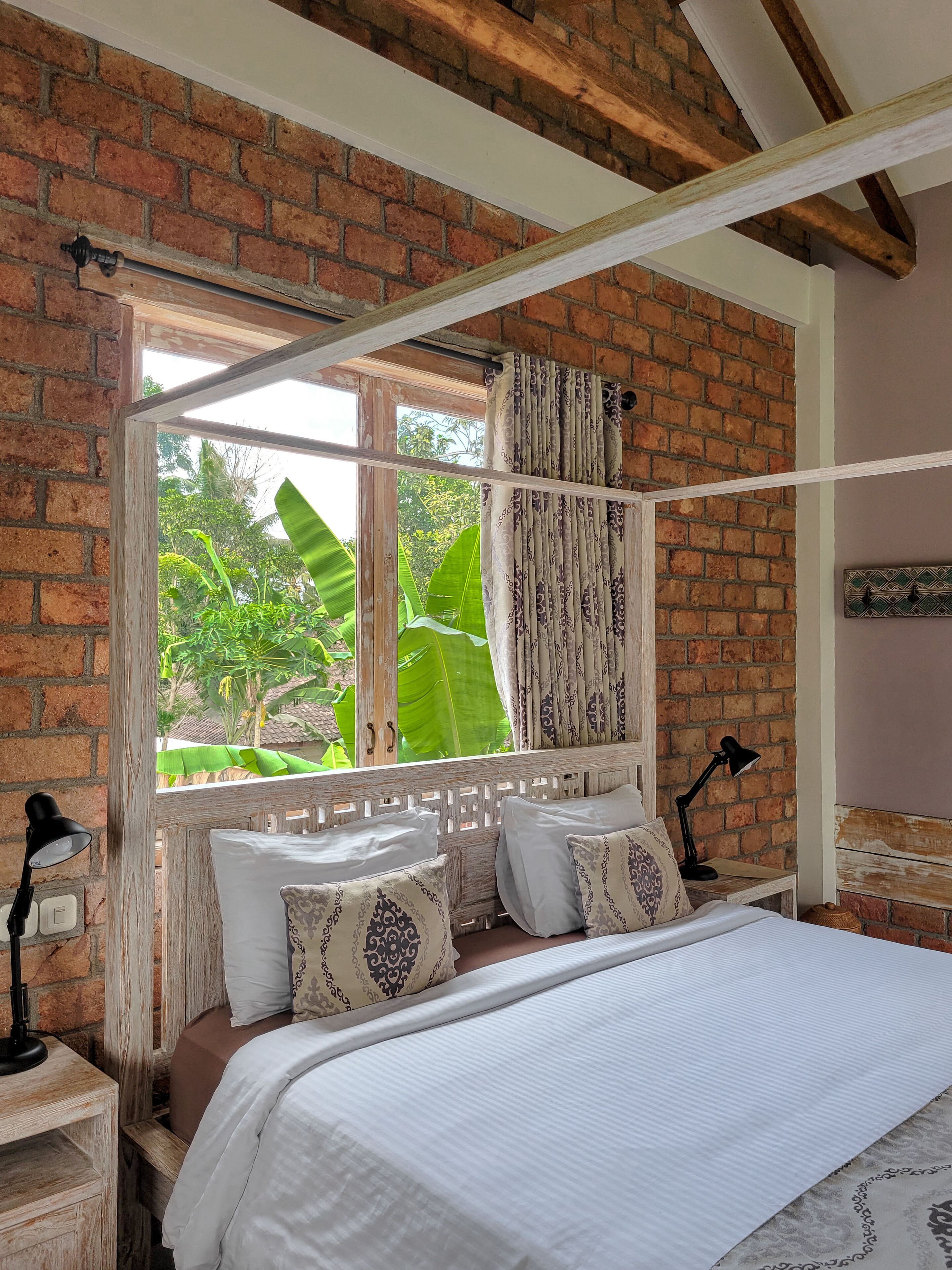 A bedroom with a brick wall, a window with curtains, a bed with patterned pillows, and a nightstand with a lamp.