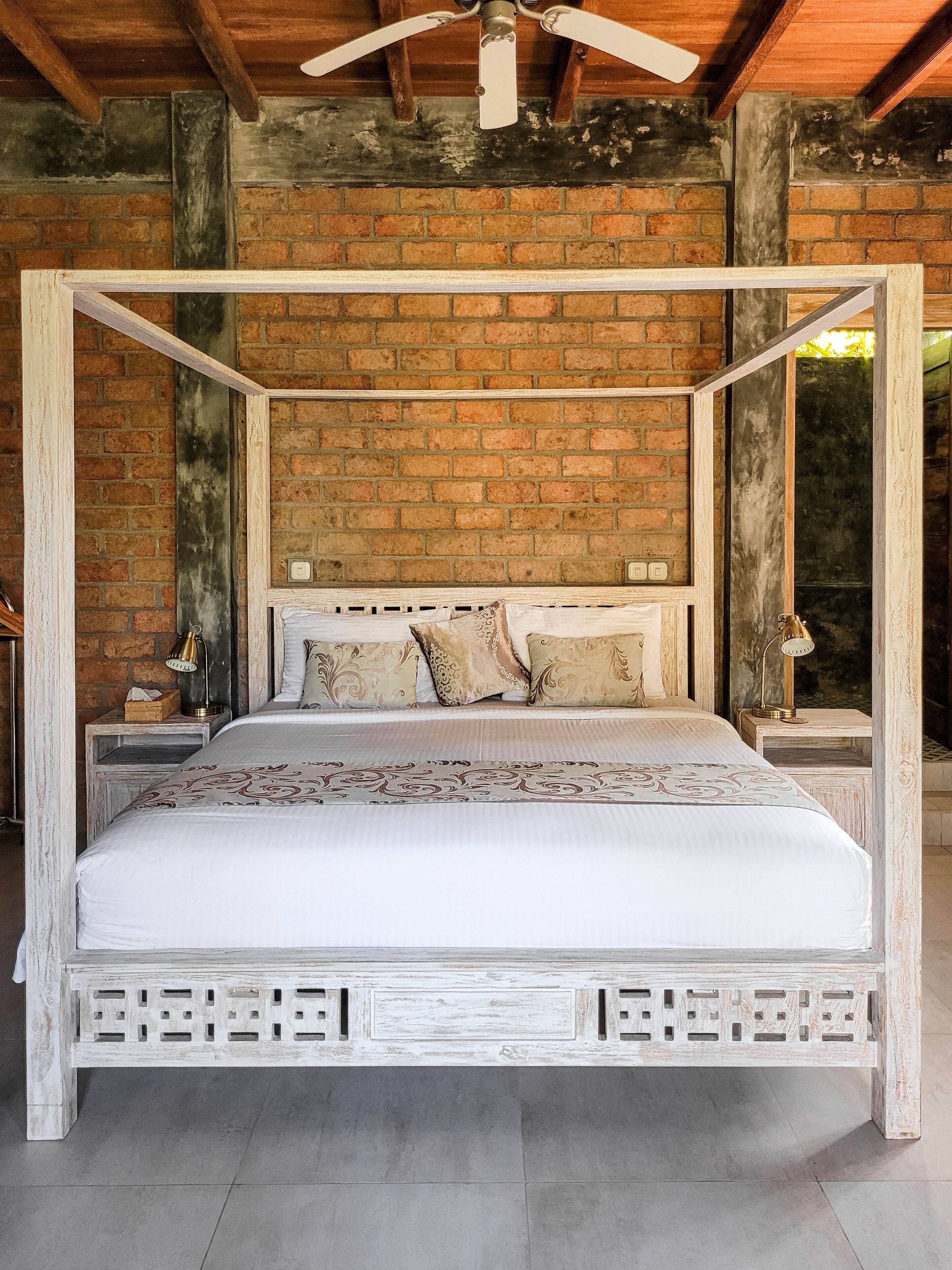 Una cama con dosel de estructura blanca y ropa de cama estampada, frente a una pared de ladrillo con vigas a la vista y un ventilador de techo.
