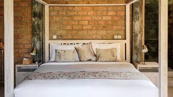 Una cama con dosel de estructura blanca y ropa de cama estampada, frente a una pared de ladrillo con vigas a la vista y un ventilador de techo.